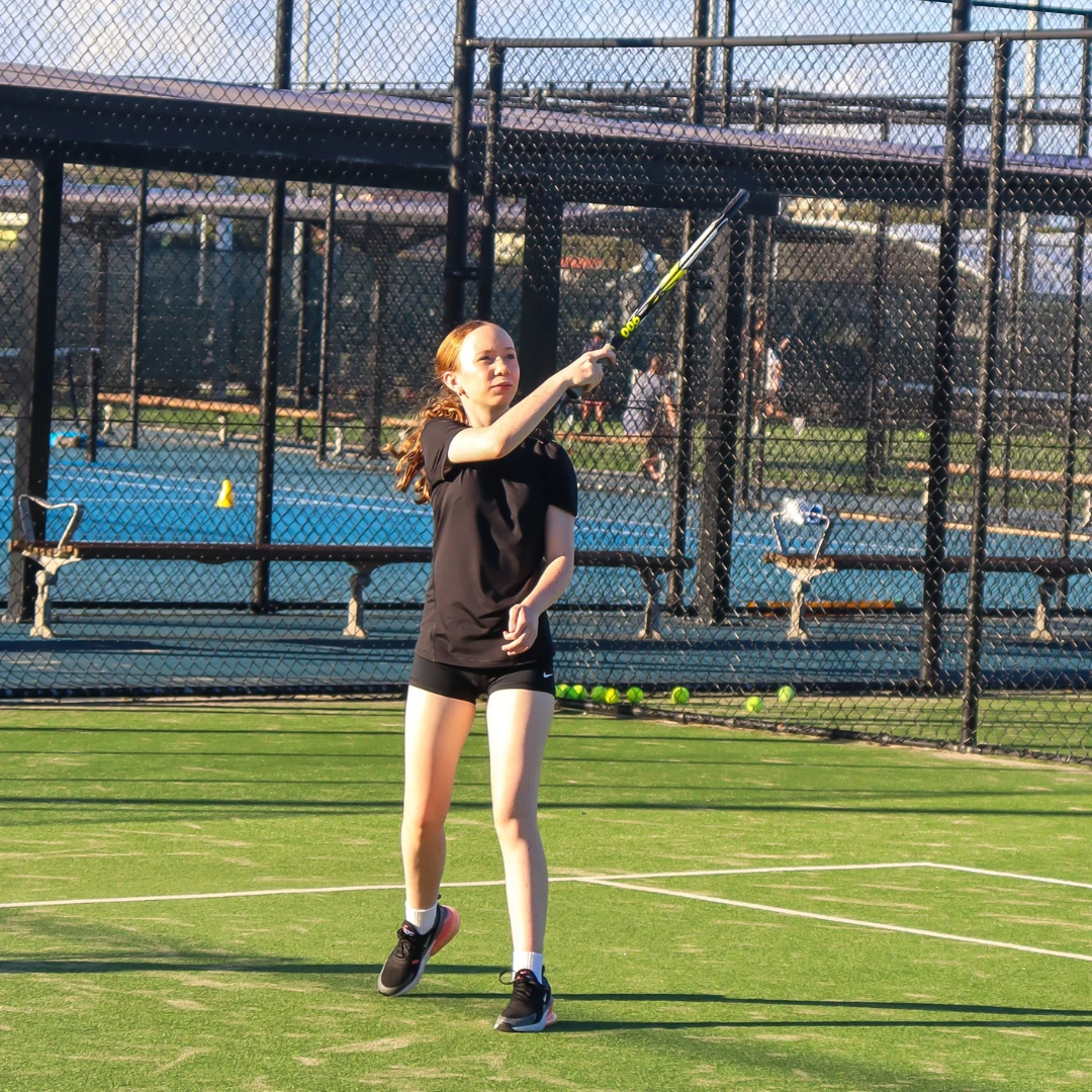 A girl on an outdoor tennis court swinging a tennis racket to hit a tennis ball.